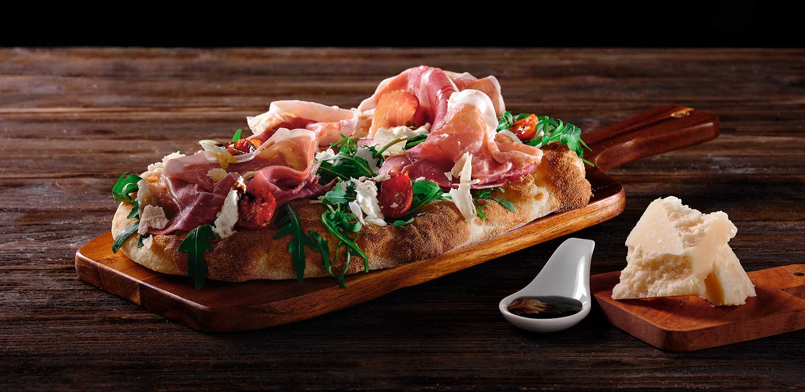 Pinsa con rucola, prosciutto crudo, pomodorini confit, parmigiano, glassa di aceto balsamico e olio evo