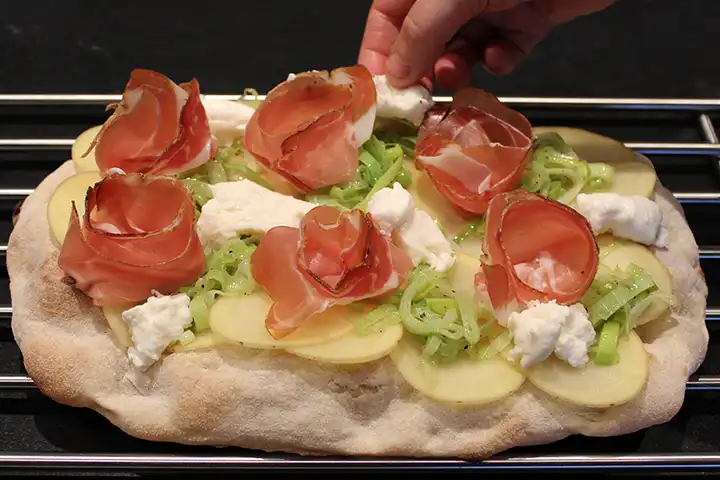 Platziere die Pinsa-Basis auf einem Rost und füge die Kartoffeln, Lauch und Speck