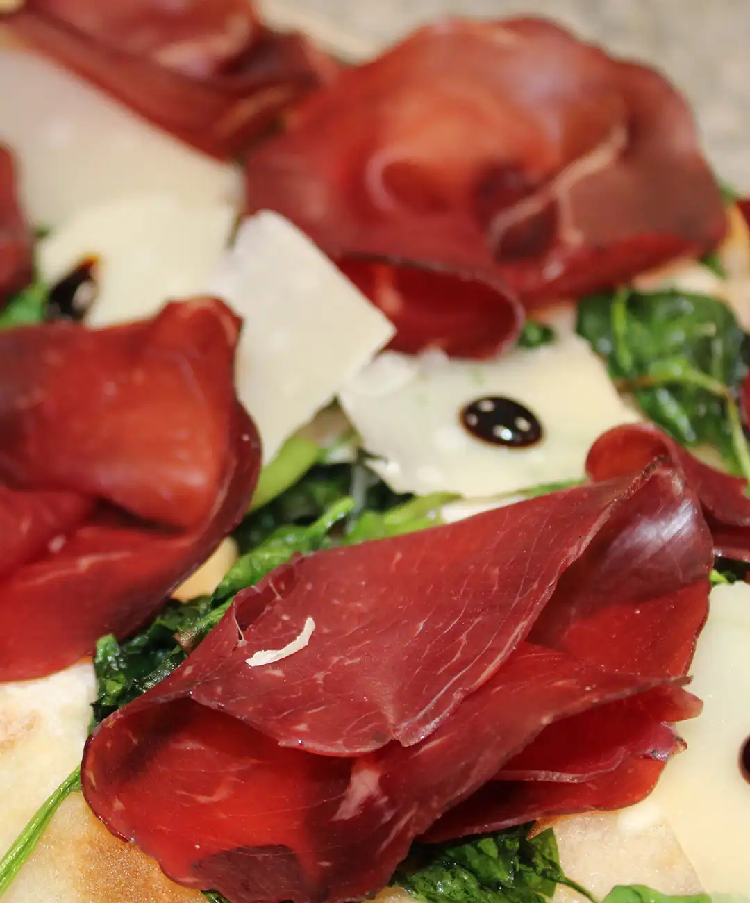 Nach dem Backen belege die Pinsa mit den Scheiben Bresaola, Parmesanspänen und ein paar Tropfen Balsamico-Essig