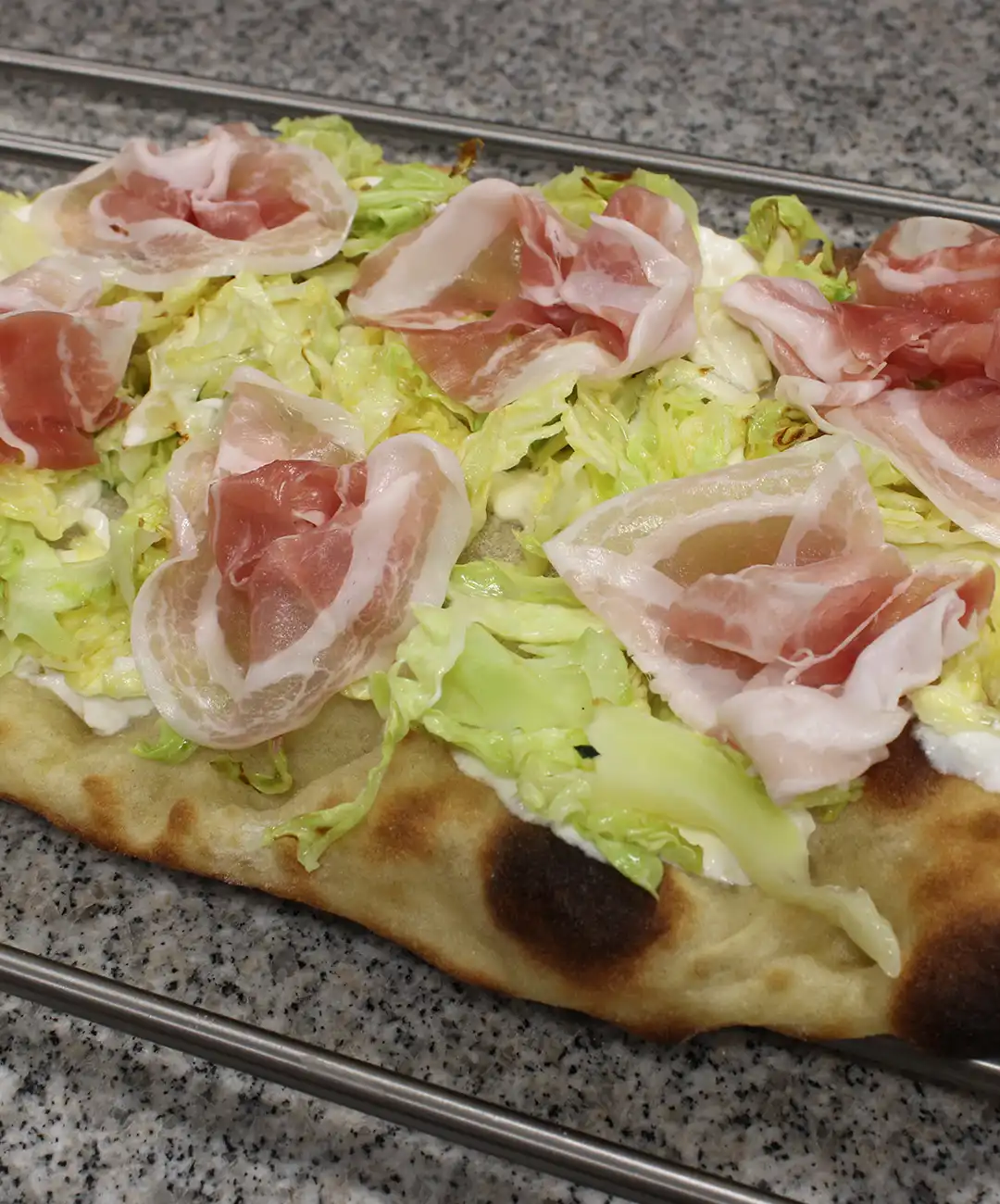 Verteile den zuvor geschnittenen Wirsing auf der Pinsa-Basis und füge die Pancetta-Scheiben hinzu.